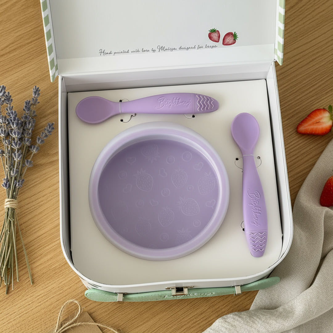 Purple children's bowl and spoons set in a box on a wooden surface with strawberries and lavender.