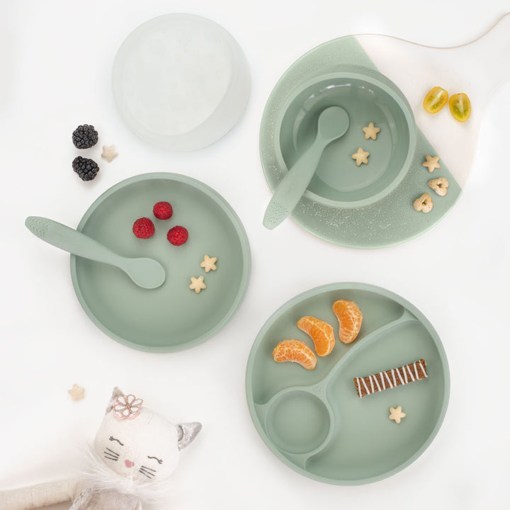 Set of green children's plates, bowls, and spoons with food on a white background