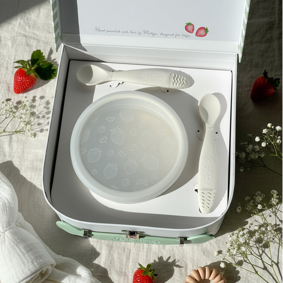 White baby bowl and spoons in a box with strawberries and flowers on a light background