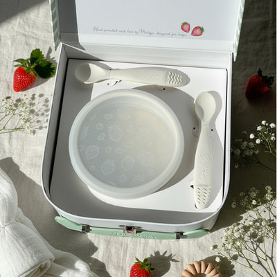 White baby bowl and spoons in a box with strawberries and flowers on a light background