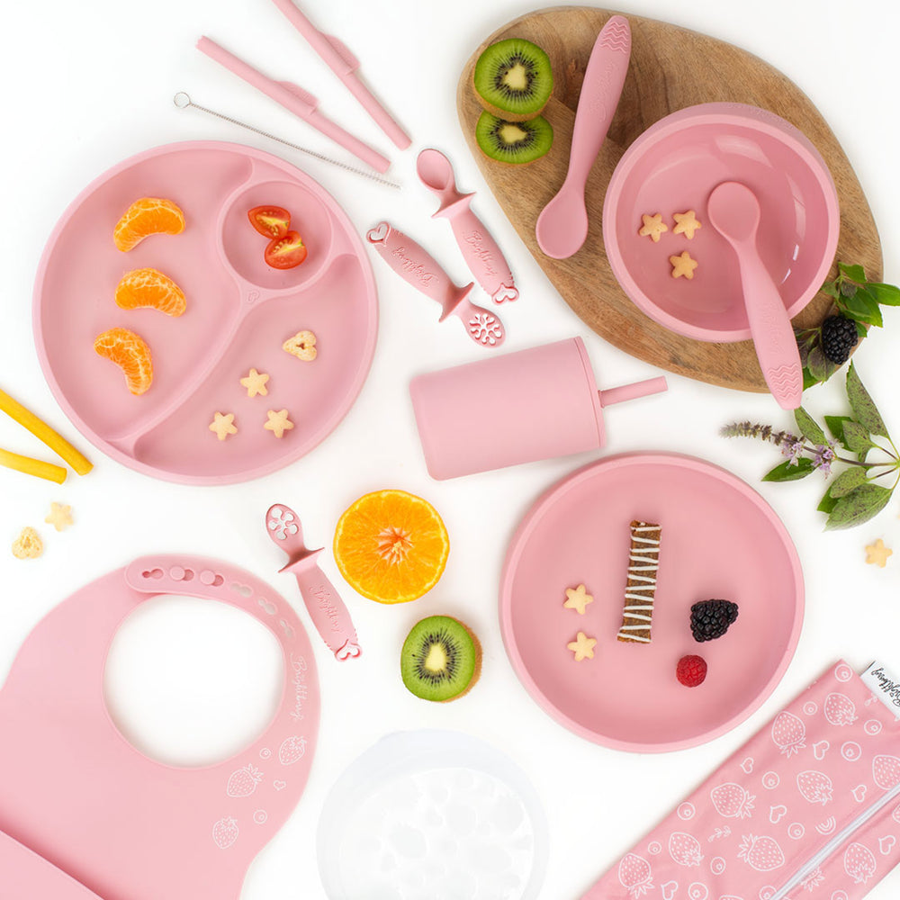 Flat lay image of a coral pink silicone feeding set for kids, including divided plates, bowls, spoons, dipping spoons, a smoothie cup with a lid, a bib, and a straw cleaning brush, surrounded by fruit slices and snacks, with a light wooden board underneath and a white background.