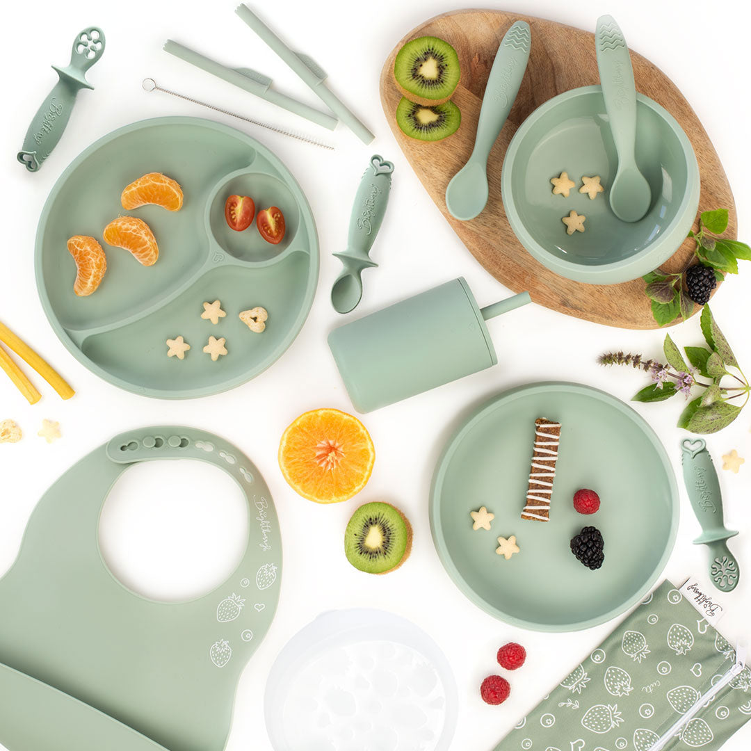 Flat lay image of a sage green silicone feeding set for kids, including divided plates, bowls, spoons, dipping spoons, a smoothie cup with a lid, a bib, and a straw cleaning brush, surrounded by fruit slices and snacks, with a light wooden board underneath and a white background.