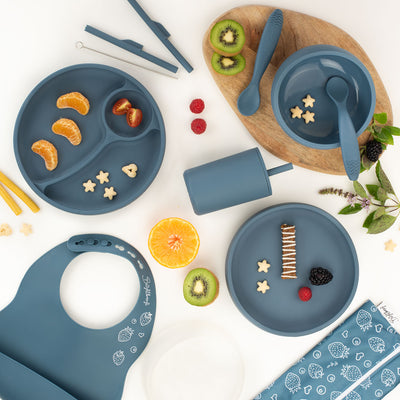Flat lay image of a navy silicone feeding set for kids, including divided plates, bowls, spoons, a smoothie cup with a lid, a bib, and a straw cleaning brush, surrounded by fruit slices and snacks, with a light wooden board underneath and a white background.