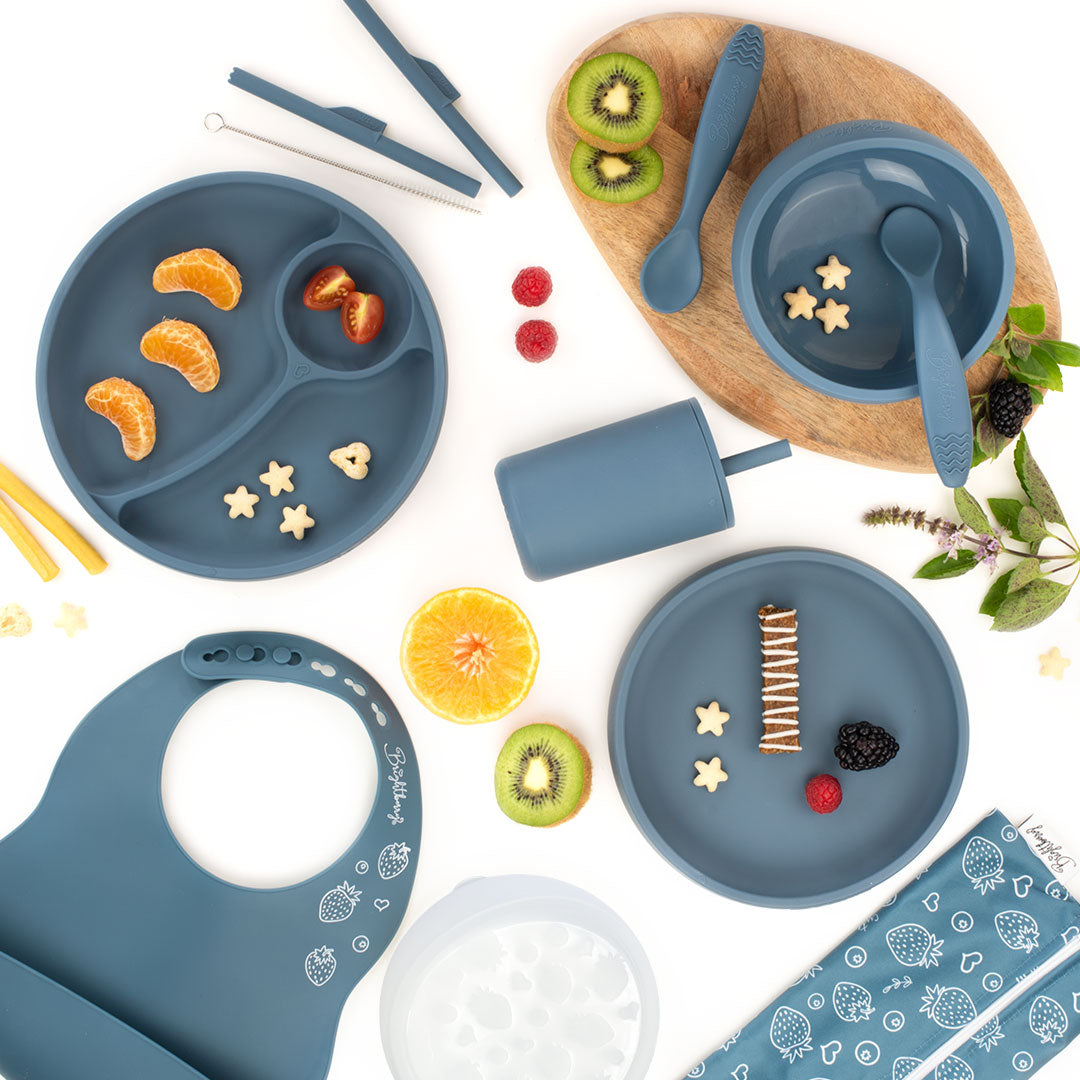 Flat lay image of a blueberry blue silicone feeding set for kids, including divided plates, bowls, spoons, a smoothie cup with a lid, a bib, and a straw cleaning brush, surrounded by fruit slices and snacks, with a light wooden board underneath and a white background.