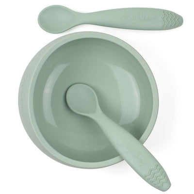 A sage-green silicone suction bowl set by Brightberry for babies. The bowl has a strong suction base that keeps it in place during mealtime, even for active toddlers. The set also includes two teething spoons in matching sage-green color. The spoons have a soft, flexible silicone tip that is gentle on gums. The bowl and spoons are BPA-free and dishwasher safe.