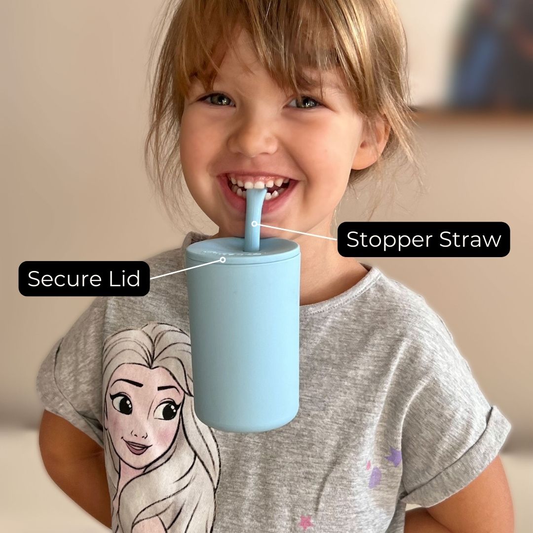 A cheerful young child with a bright smile is holding a light blue Brightberry smoothie cup. The cup is featured with a 'Secure Lid' and a 'Stopper Straw' as labeled in the image, emphasizing its child-friendly and mess-preventing design. The child's gray T-shirt features a graphic of a Disney's Elsa, adding a playful touch to the photo.
