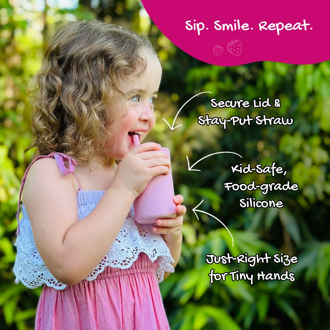 A young girl smiles while sipping from a pink Brightberry Smoothie Cup outdoors. The cup features a secure lid with a stay-put straw. Text on the image highlights key features: "Secure Lid & Stay-Put Straw," "Kid-Safe, Food-Grade Silicone," and "Just-Right Size for Tiny Hands." The caption "Sip. Smile. Repeat." appears at the top in a magenta banner.