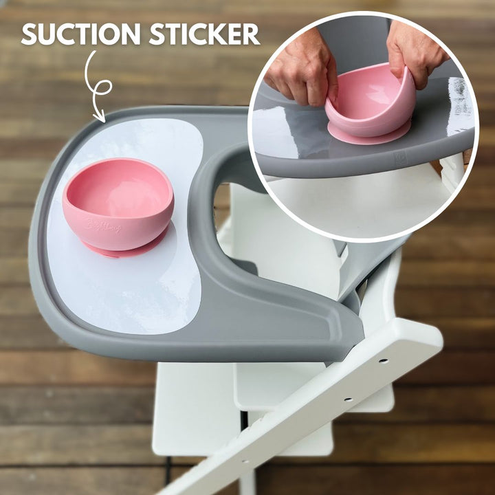 Image featuring a Brightberry suction sticker on a grey Stokke Tripp Trapp high chair tray. The main image shows a pink Brightberry bowl securely attached to the sticker. An inset close-up on the right highlights a person’s hands pulling the bowl off the suction sticker, demonstrating its effectiveness. Text at the top reads 'SUCTION STICKER'.