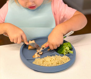 toddler girl with silicone bib eating independently using kids utensils and suction baby plate in blueberry blue colour