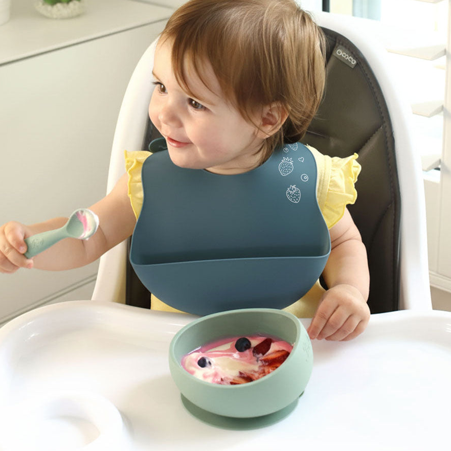 Child in a high chair with a blue bib, green bowl, and spoon, sitting at a dining table.