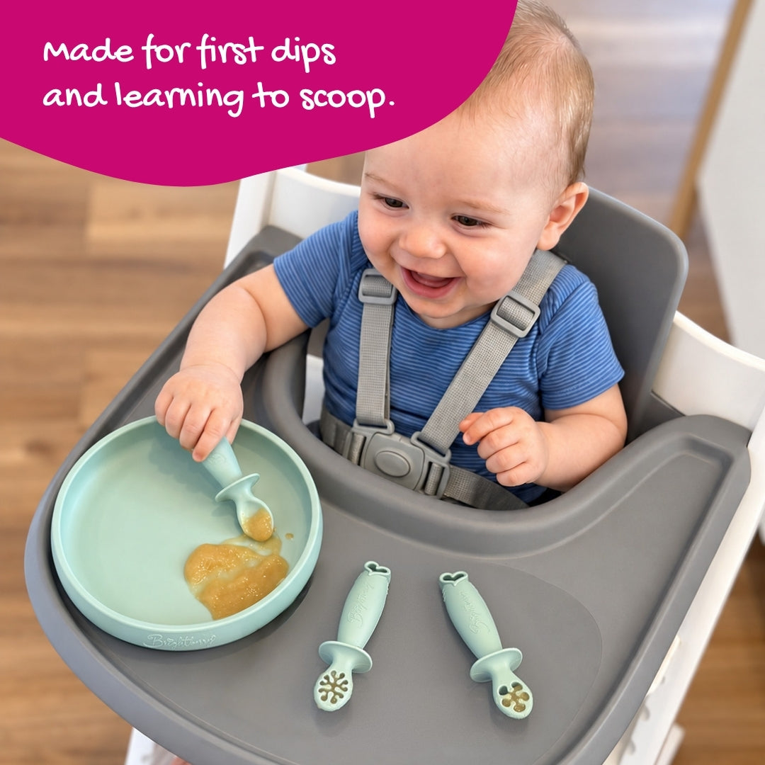 Baby holding a Sage theFirst spoon in a plate of purée, with two dipping spoons on the highchair tray.