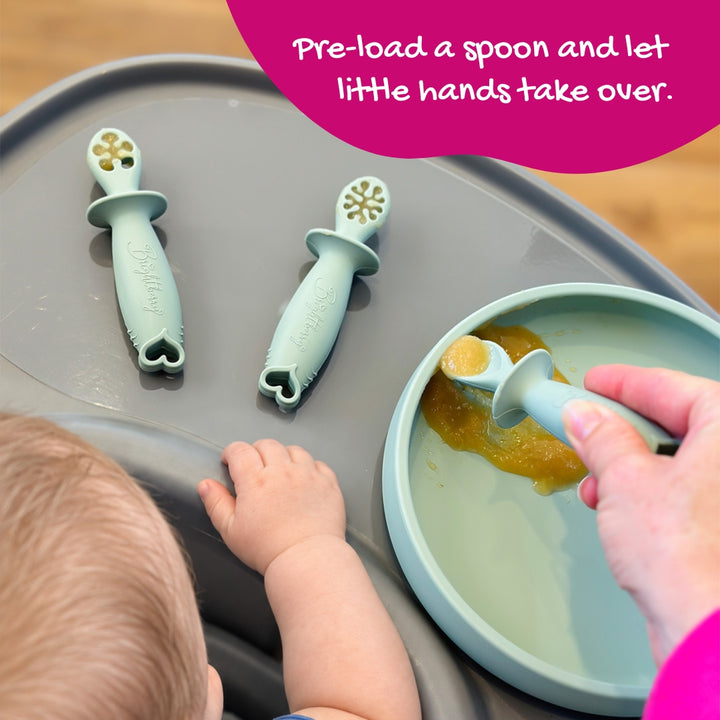 Parent loading baby spoon with purée while baby reaches towards two dipping spoons on the tray in sage colour.