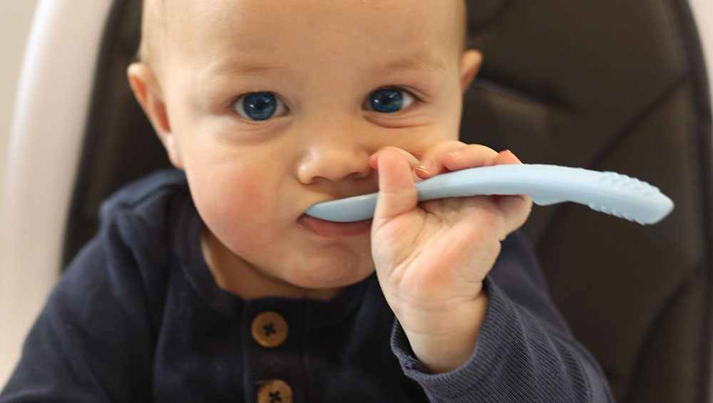 Baby holding a blue silicone spoon in a high chair