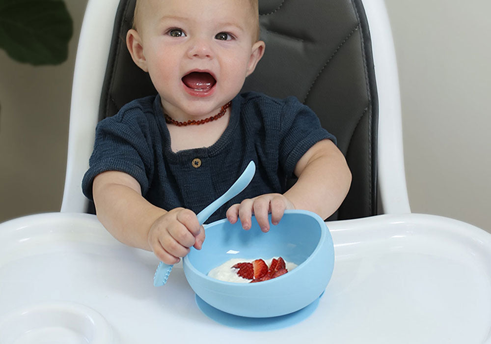 toddler boy holding silicone spoon trying to unstick suction bowl from the high chair tray