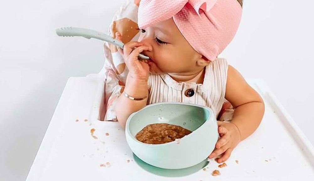 baby girl self feeding baby food with Brightberry silicone spoon and suction bowl in sage colour siting in a high chair