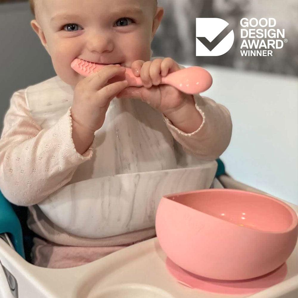 A coral-colored silicone suction bowl set with two teething spoons designed for babies, by Brightberry. The bowl features a suction base to keep it in place during mealtime.