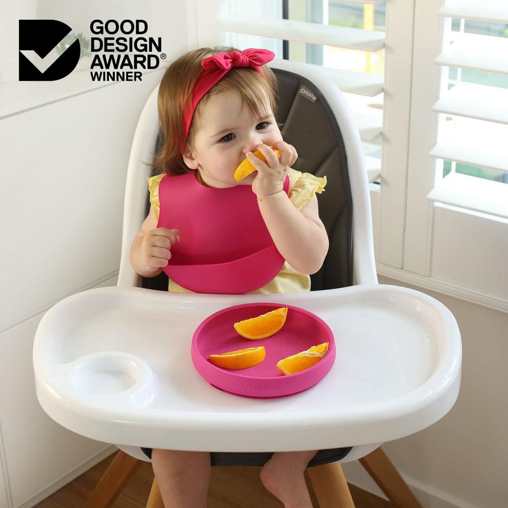 Toddler girl sitting in a high chair eating an orange from a silicone suction plate in hot pink colour. She is wearing a fuchsia pink silicone bib